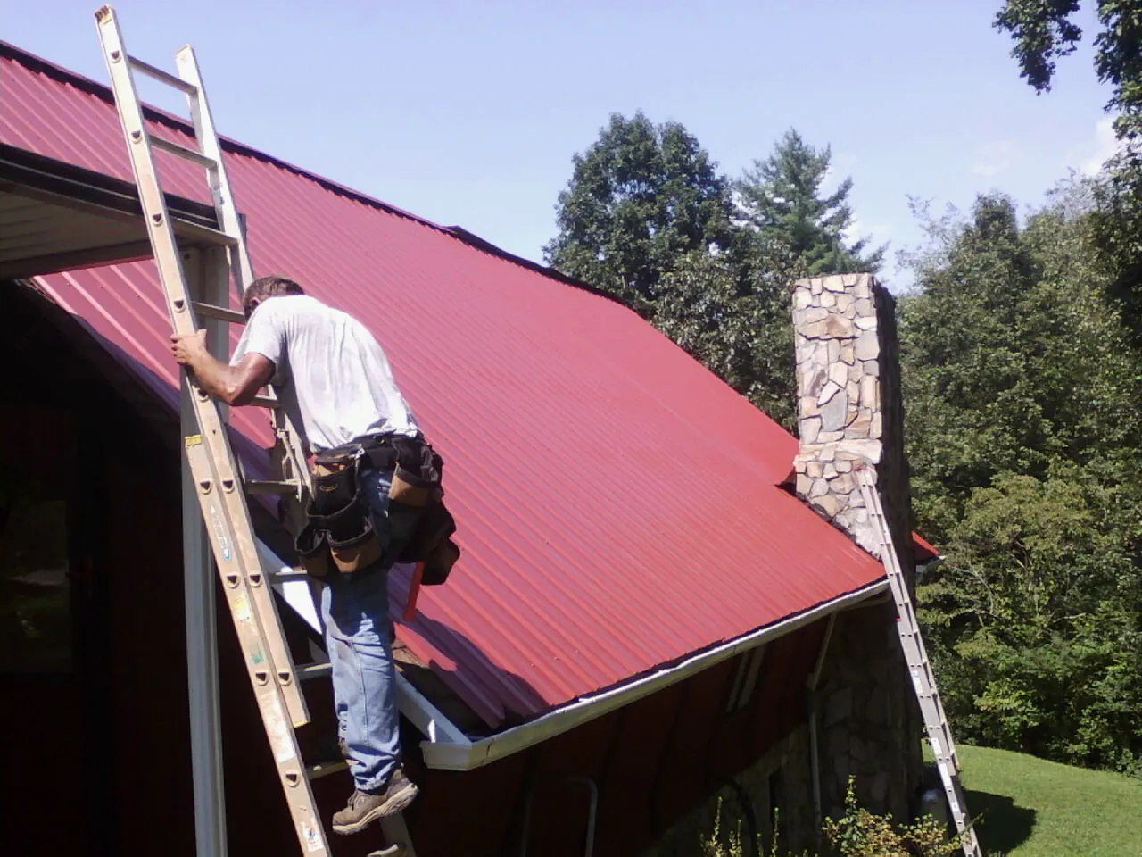Roofer on a ladder beside a red metal roof and stone chimney — Soffit & Fascia Repair by Roofing Brothers in Bruno